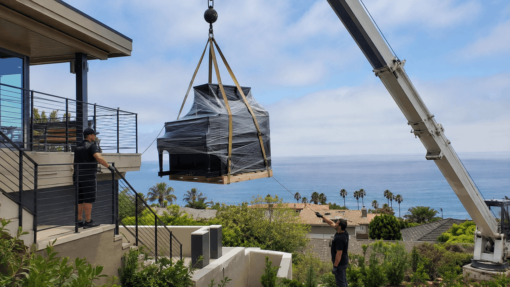 Crane lifting wrapped grand piano to luxury home balcony overlooking ocean Los Angeles