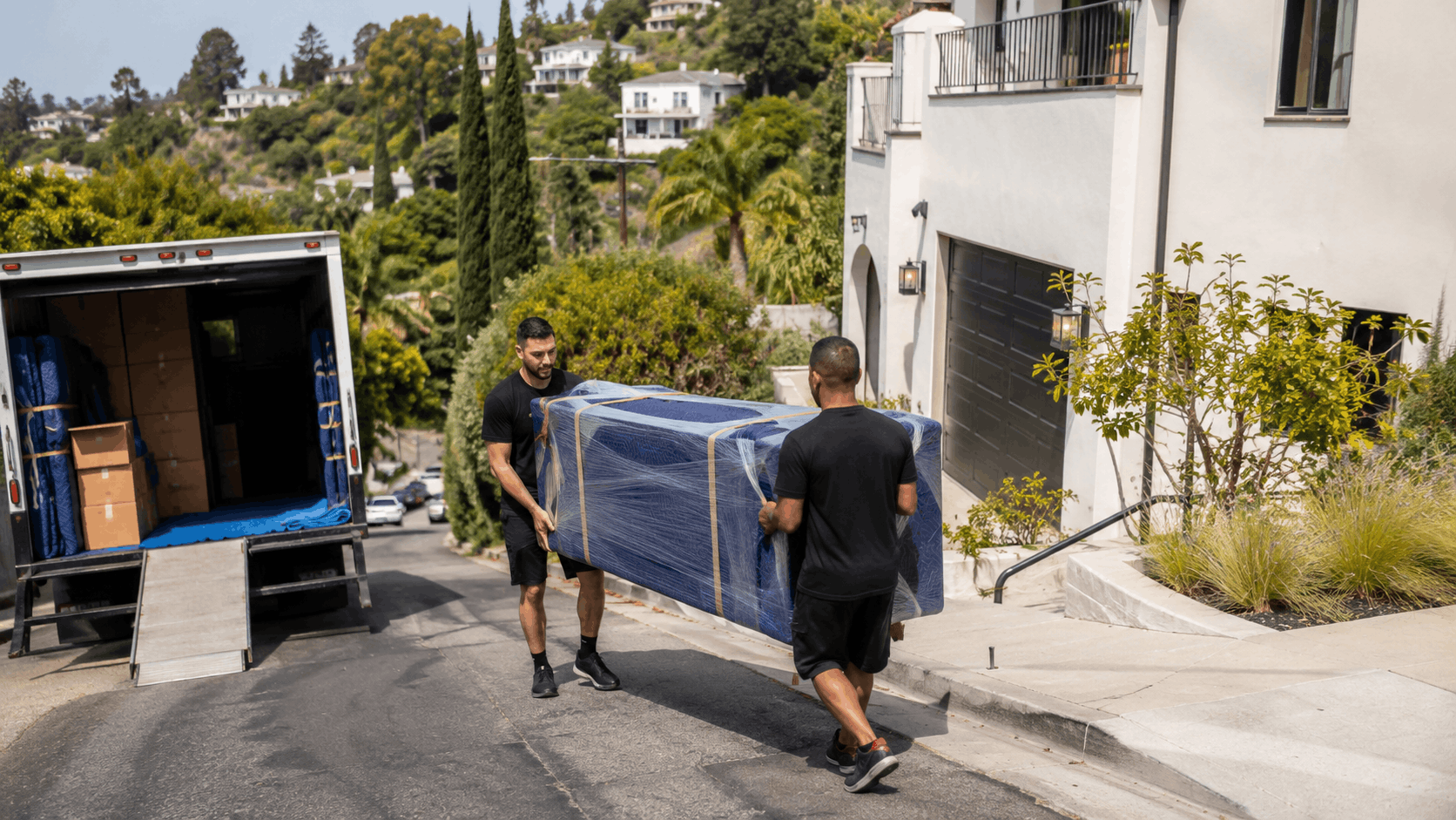 Two movers carrying wrapped bulky furniture from truck to hillside Los Angeles home