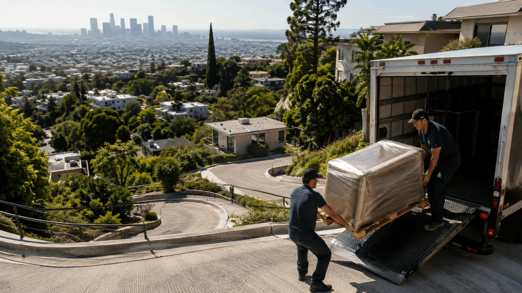 Two movers unloading wrapped furniture from truck on steep Hollywood Hills driveway overlooking Los Angeles skyline