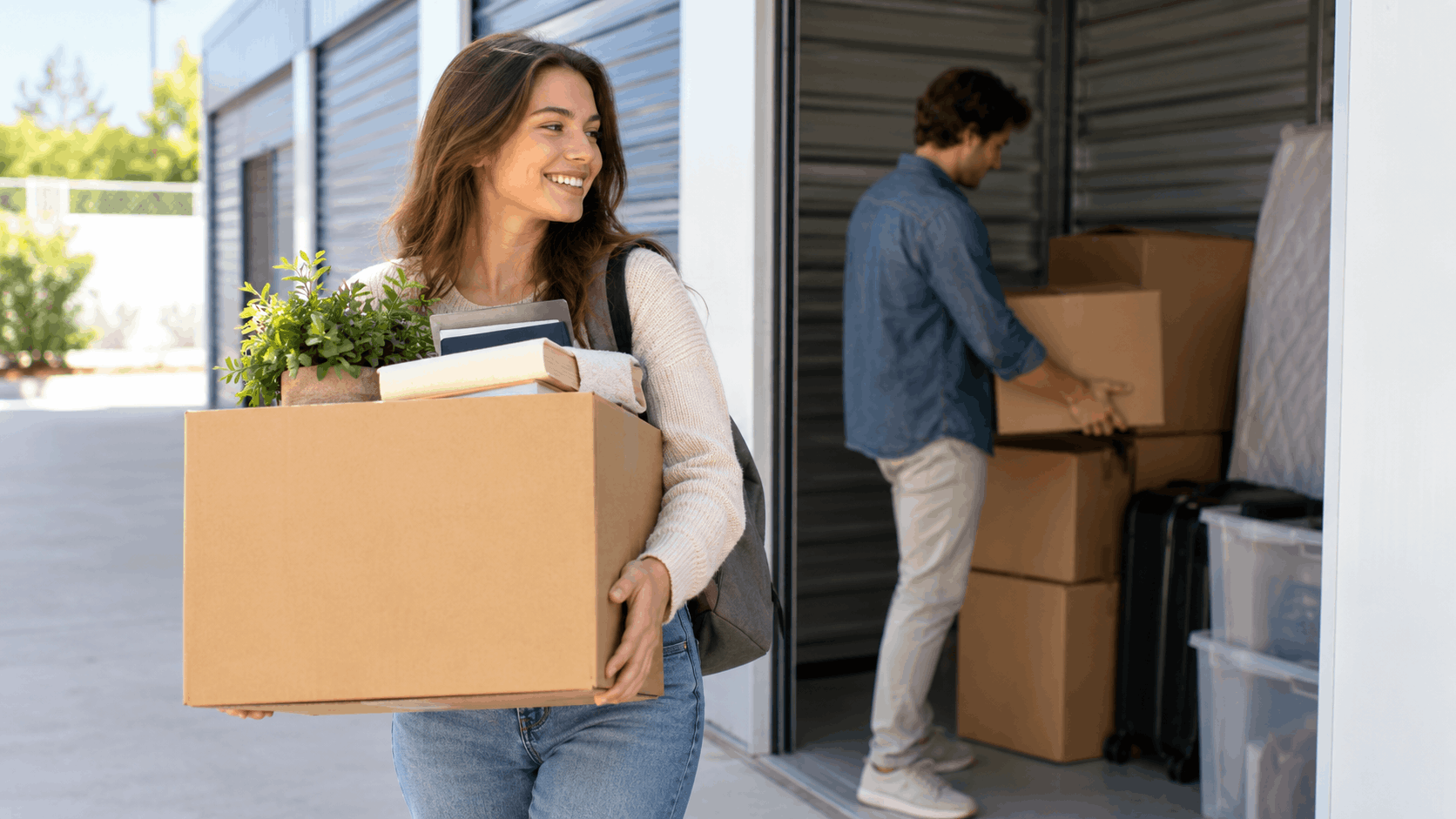 Smiling woman carrying box outside Los Angeles self-storage facility with man unloading